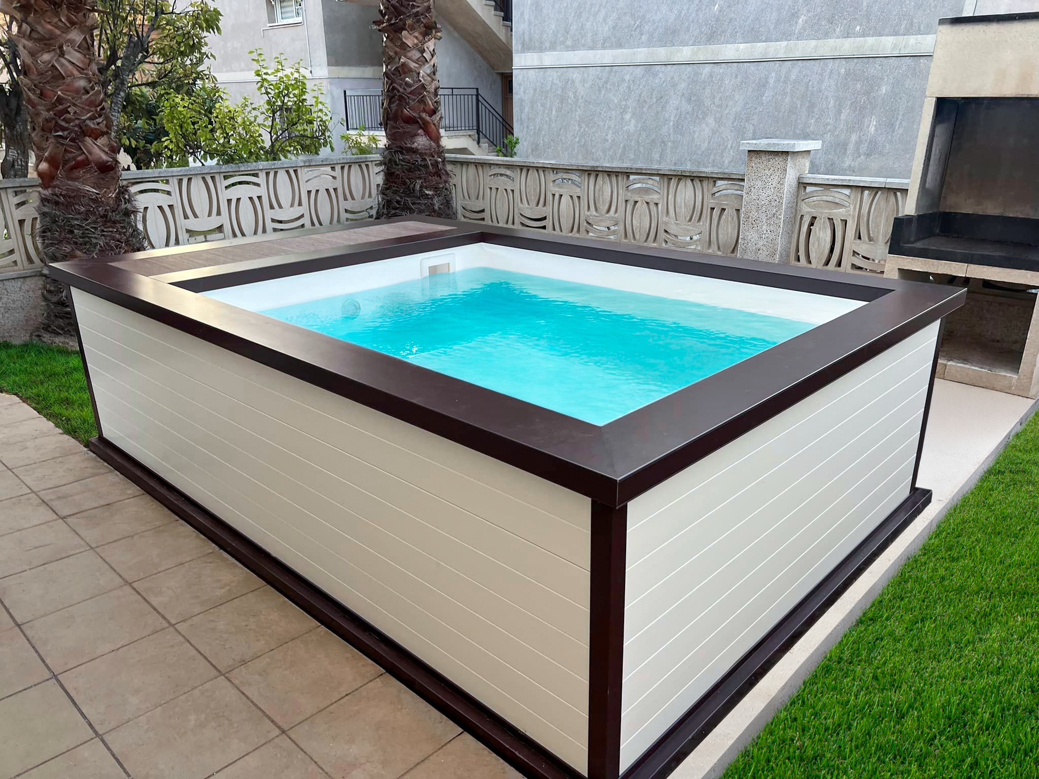 Compact above-ground aluminium pool with dark wood trim and tiled patio, nestled in a residential courtyard